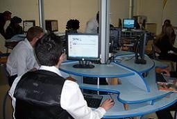 Photo salle informatique, bureaux circulaires bleus en bois, élève vu de dos face à un écran.